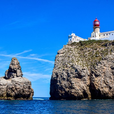Cliff with lighthouse and rock formation in ocean under clear blue sky.