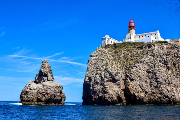 Cliff with lighthouse and rock formation in ocean under clear blue sky.