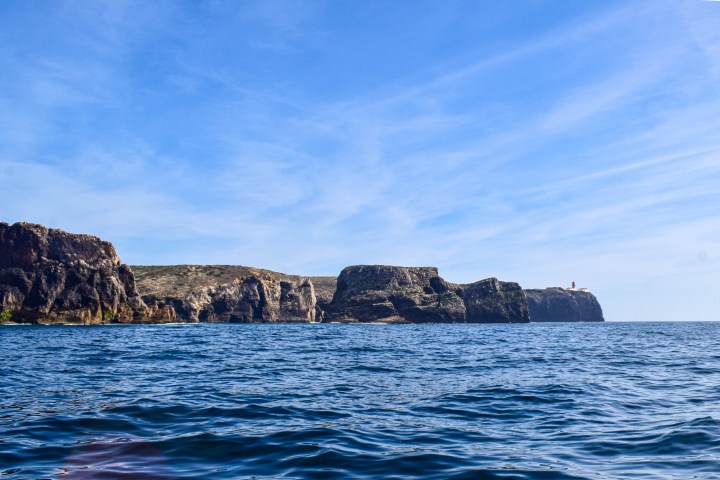 Rocky coastline with cliffs and a lighthouse under a clear blue sky.