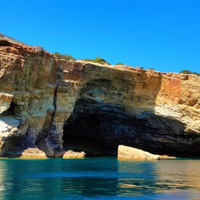 Colorful rocky cliffs with cave openings by a clear blue sea under a bright blue sky.
