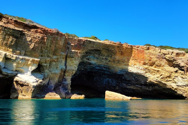 Colorful rocky cliffs with cave openings by a clear blue sea under a bright blue sky.