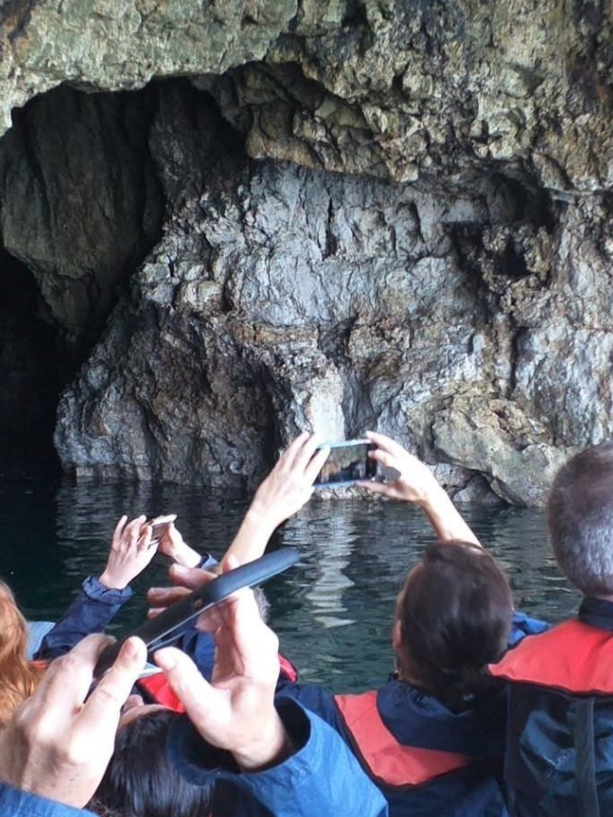 People in a boat taking photos of a rocky cave interior by the water.