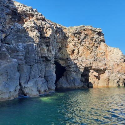 Rocky cliffside with a cave entrance by a body of water under a clear blue sky.
