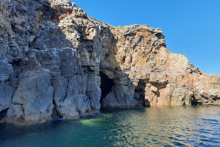 Rocky cliffside with a cave entrance by a body of water under a clear blue sky.