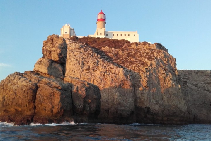 Lighthouse atop rocky cliff by the sea under clear blue sky at sunset.