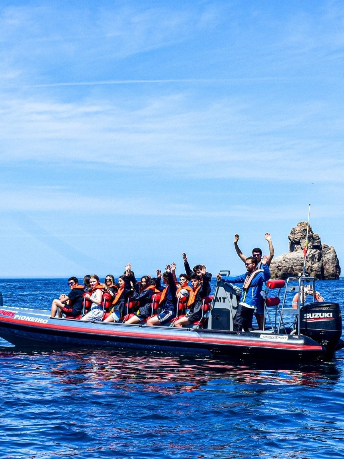 Two boats with people in life jackets in ocean near a rocky cliff and lighthouse.