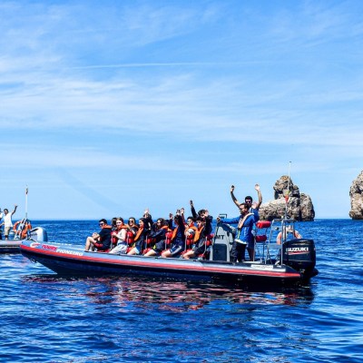 Two boats with people in life jackets in ocean near a rocky cliff and lighthouse.