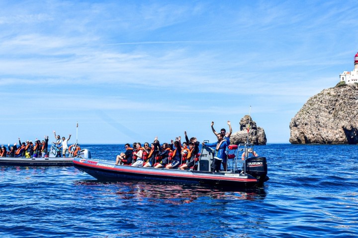 Two boats with people in life jackets in ocean near a rocky cliff and lighthouse.