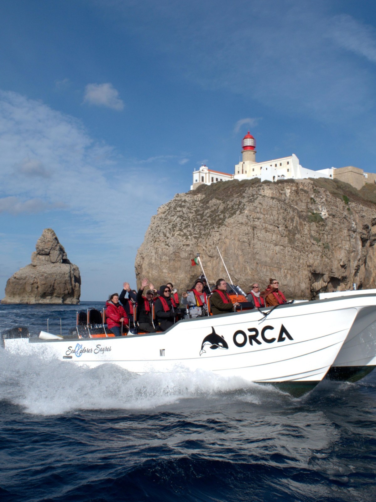 White boat named 'ORCA' with passengers in life jackets, cruising near cliffs and lighthouse on a sunny day.