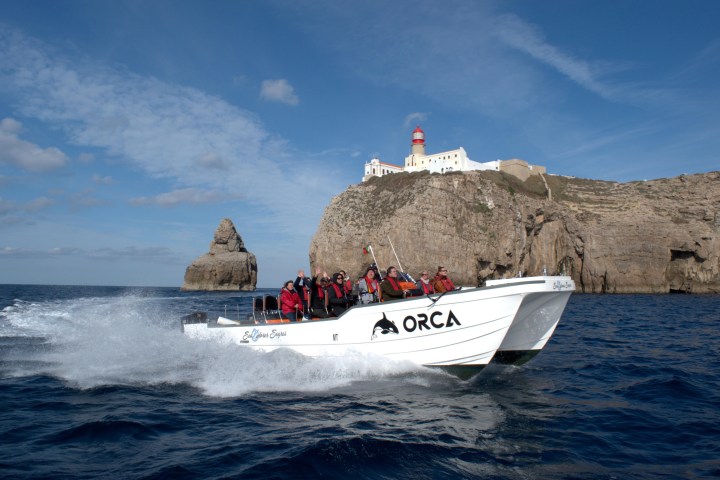 White boat named 'ORCA' with passengers in life jackets, cruising near cliffs and lighthouse on a sunny day.