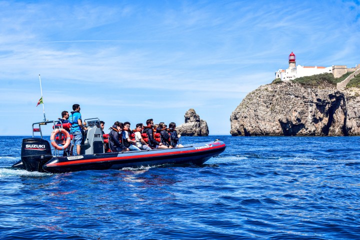 People on a boat touring near a rocky coastline with a lighthouse on top.