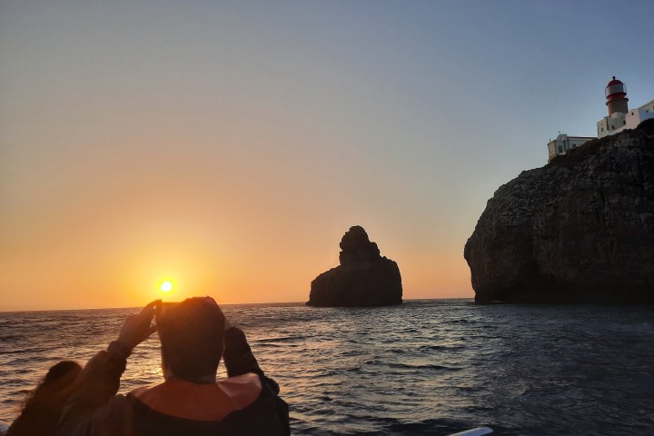 Sunset over ocean with rocky cliffs, lighthouse, and silhouetted person on a boat.