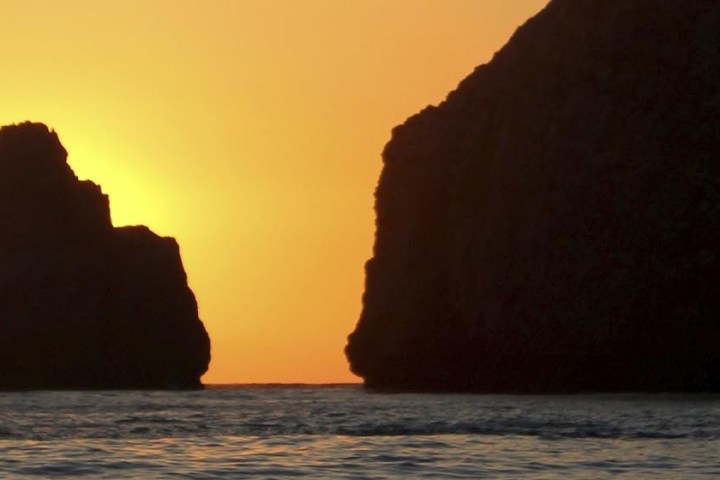Silhouette of rocks at sunset over the ocean with a glowing orange sky.