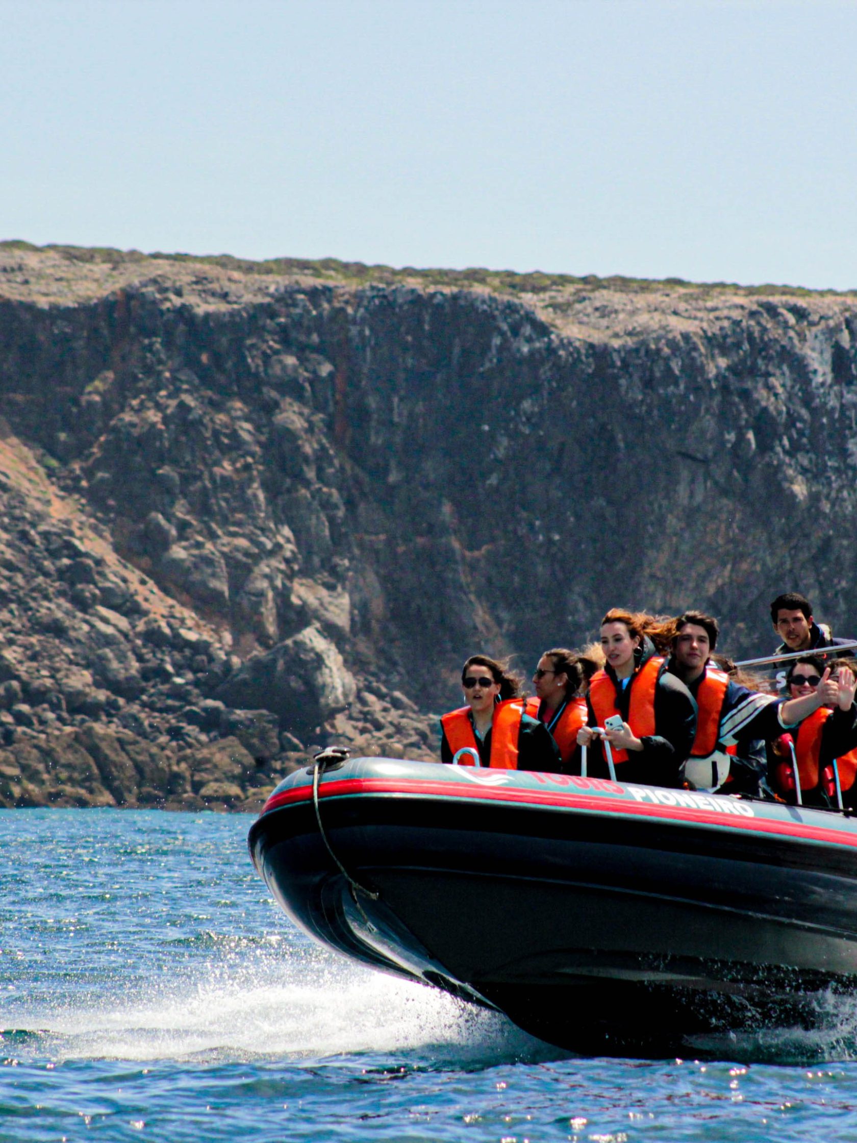 Speedboat with people wearing orange life jackets on water near a rocky coastline.