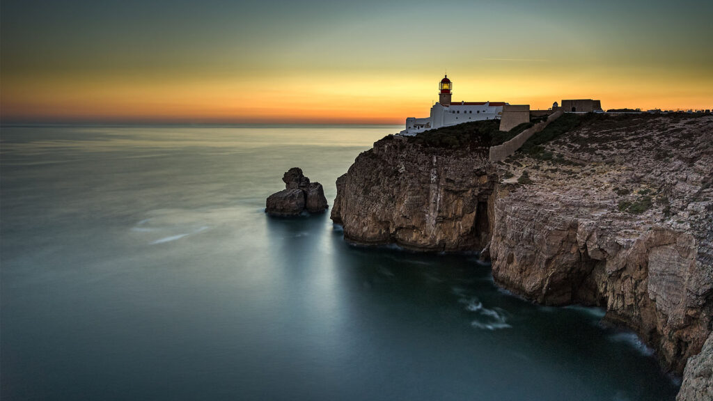 Lighthouse on a cliff at sunset over calm sea with orange sky.